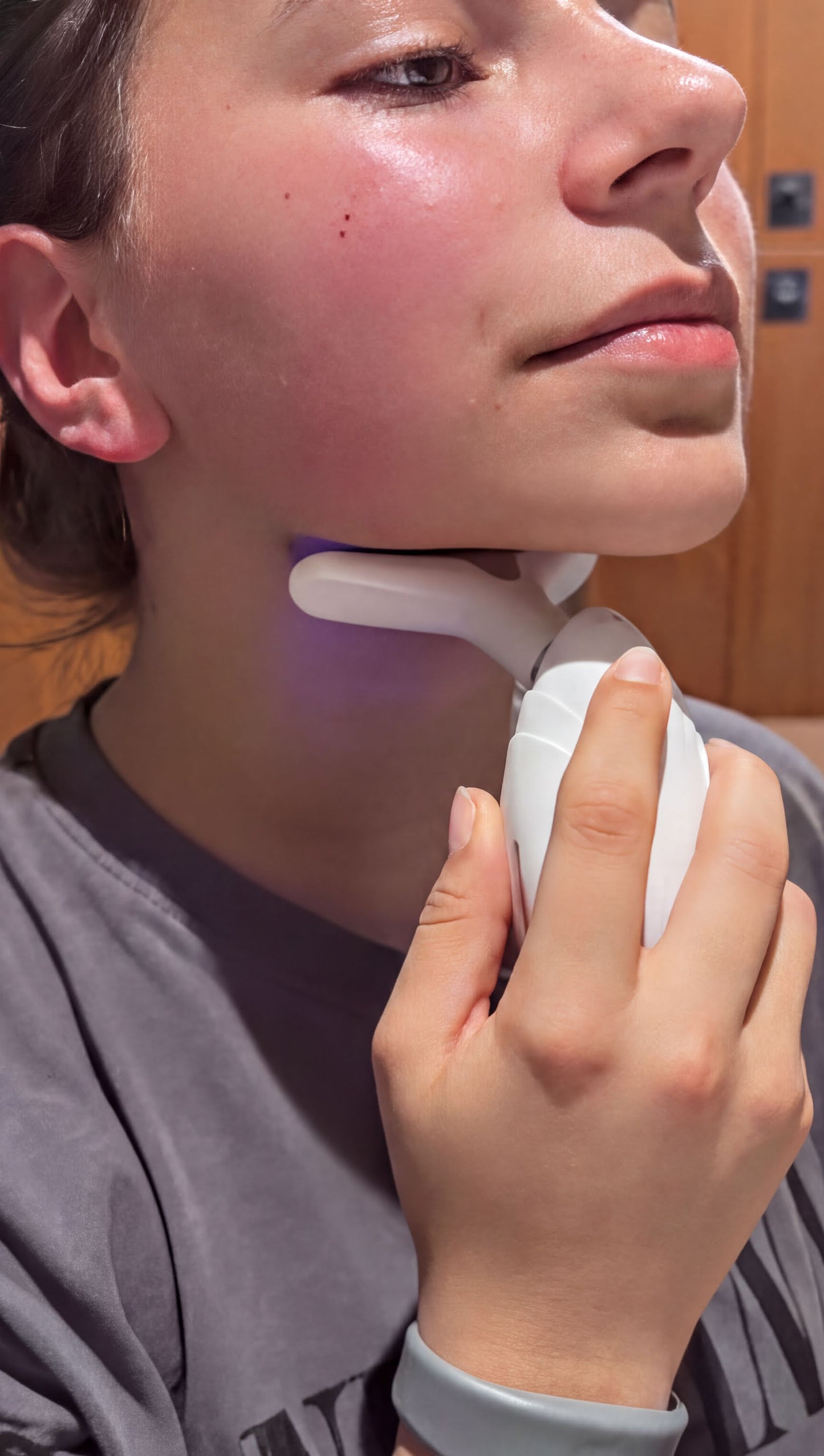 A person is holding a white device with a light on it, possibly a skin-enhancing device, and appears to be using it on their face.