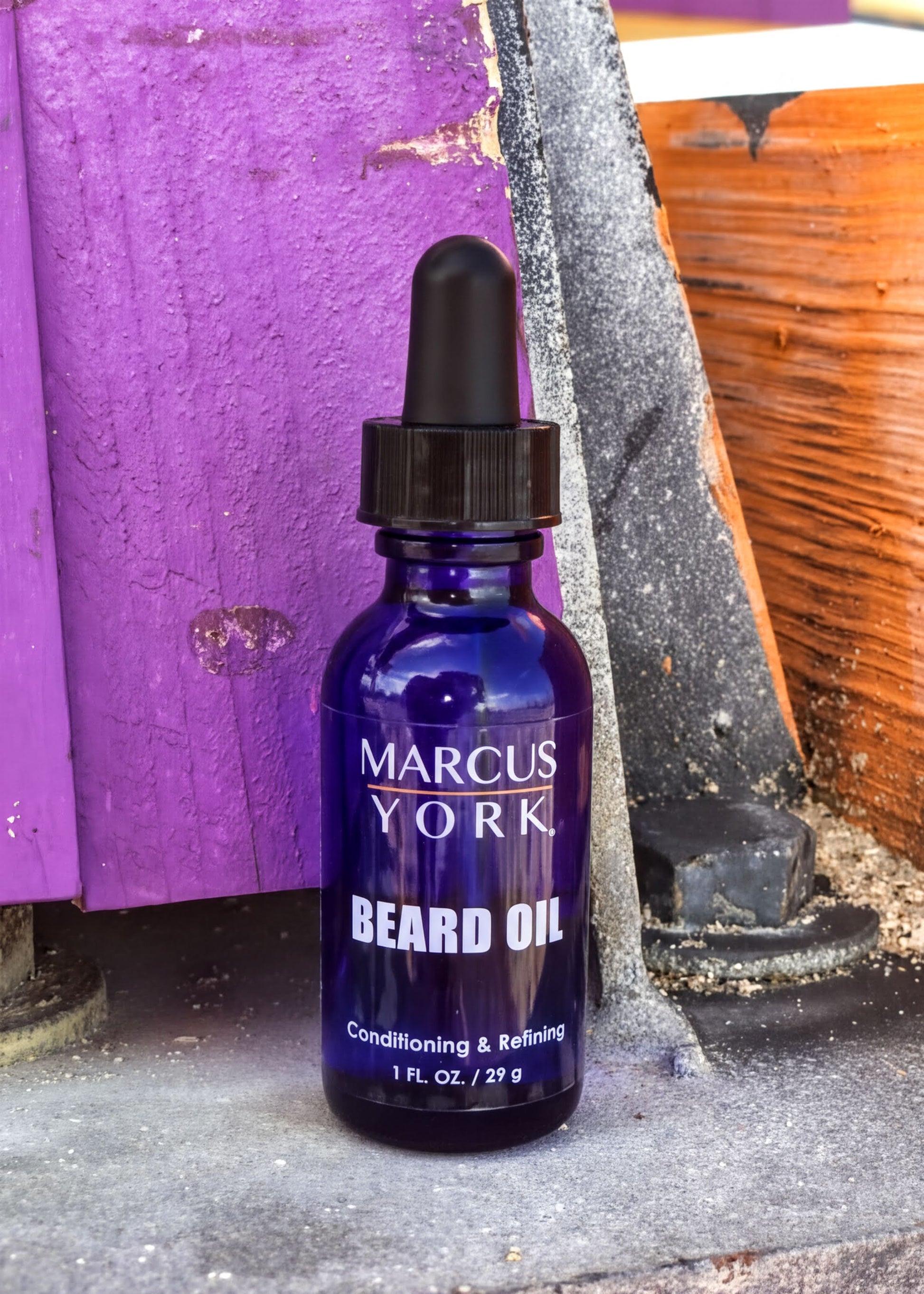 A bottle of Marcus York Beard Oil is standing on a concrete surface next to a wooden fence.