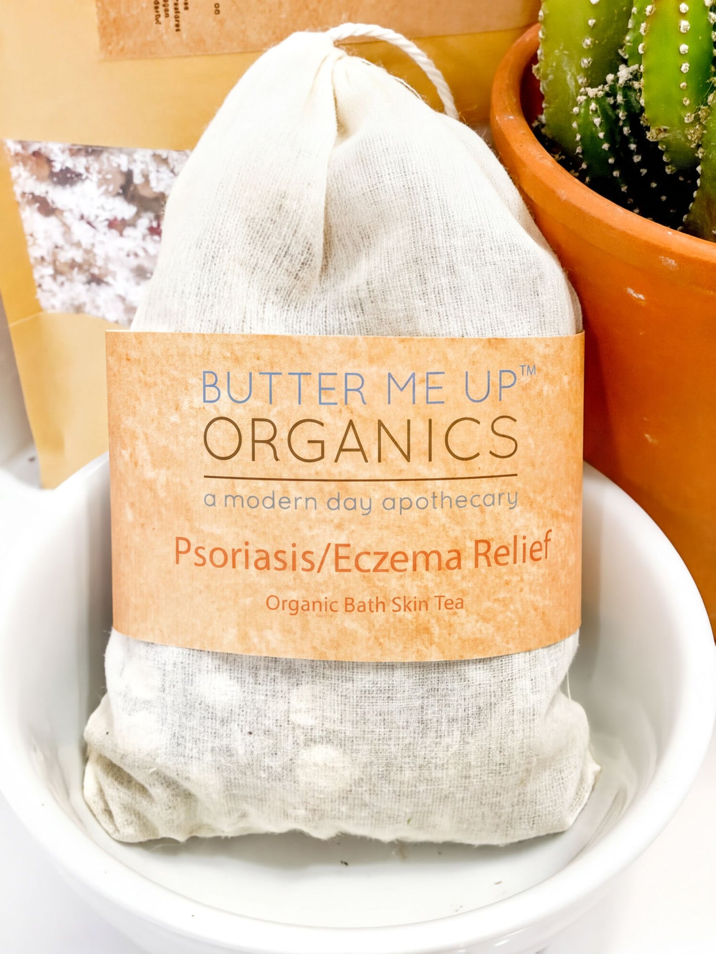 A white bowl containing a tea bag labeled "Butter Me Up ORGANICS" and "Psoriasis/ Eczema Relief Organic Bath Skin Tea".