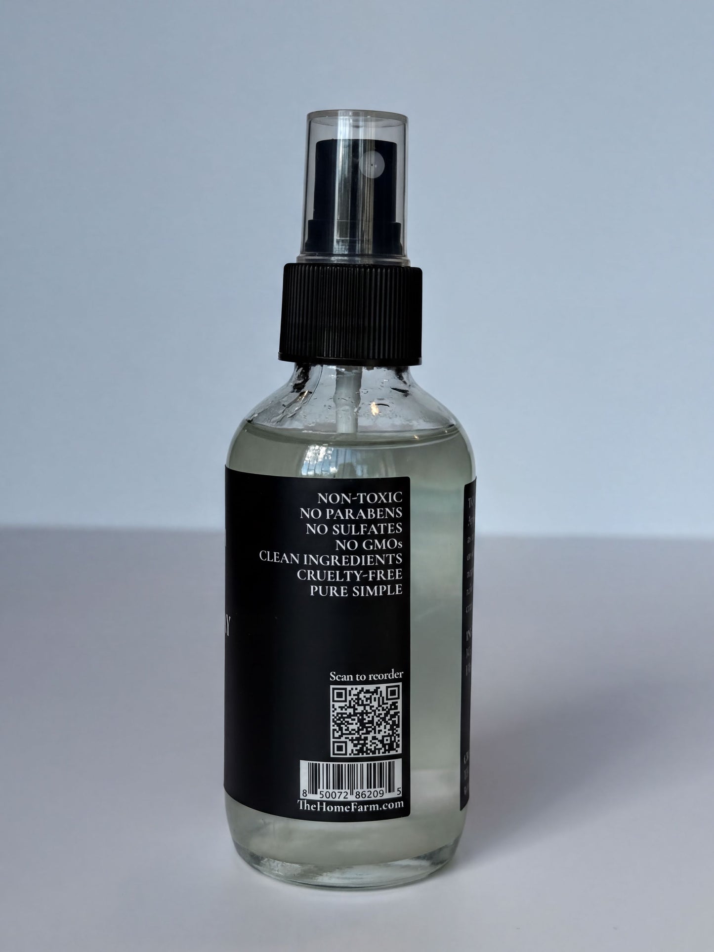 a clear glass spray bottle with a black cap, containing a clear liquid. The bottle has a label with text and a QR code.