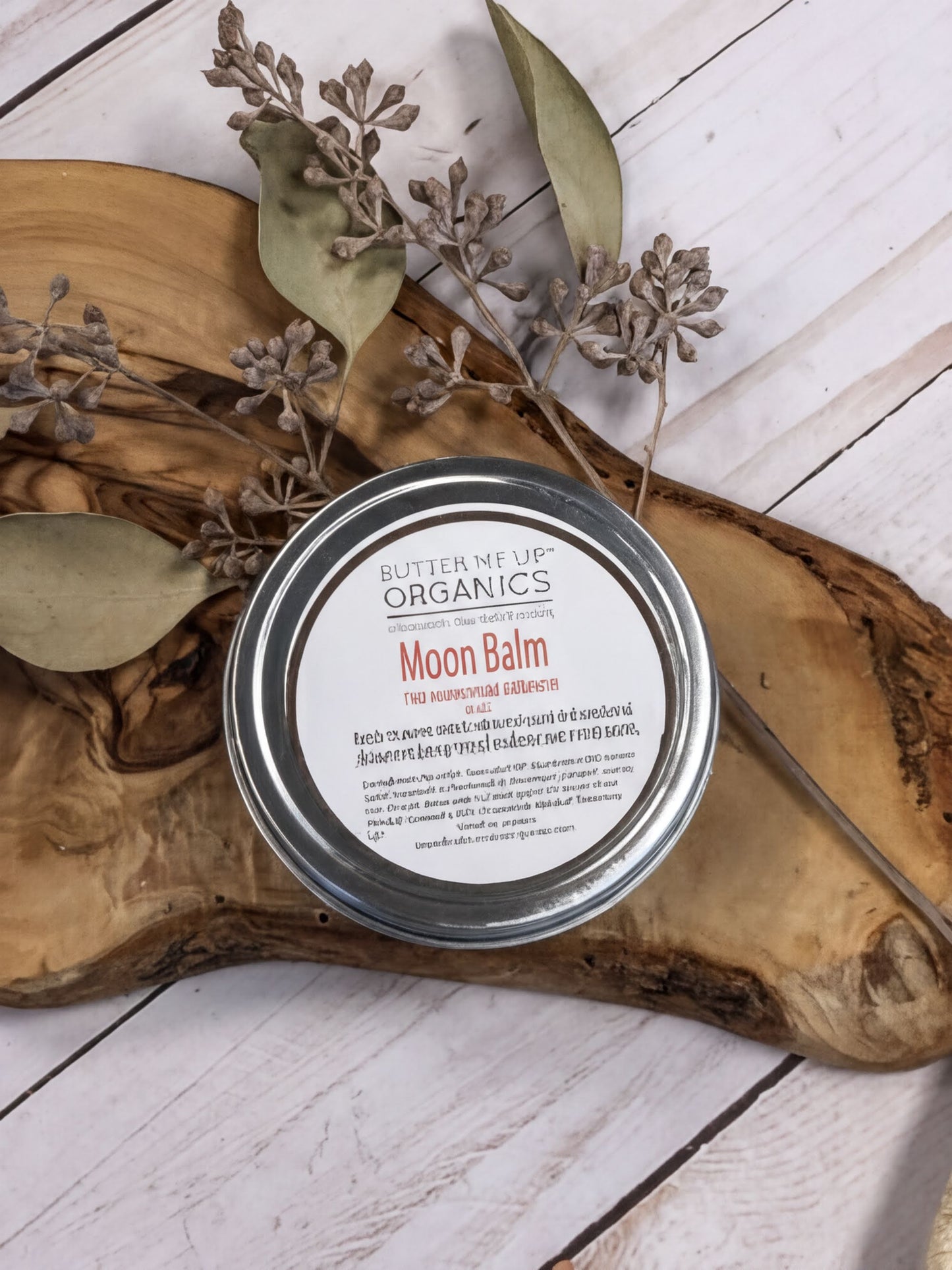 A round tin of "Moon Balm" is placed on a wooden surface, surrounded by dried leaves and branches.