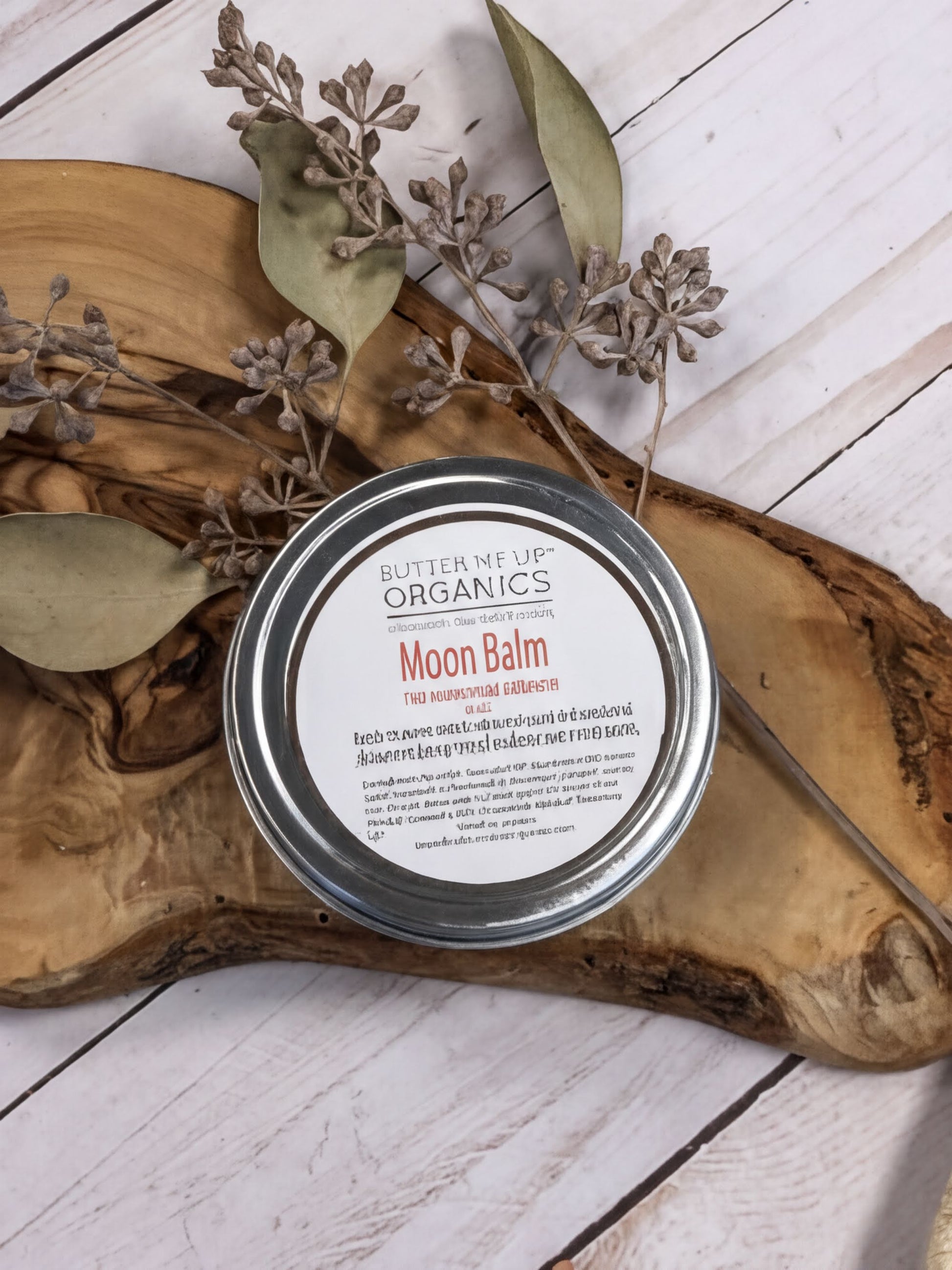 A round tin of "Moon Balm" is placed on a wooden surface, surrounded by dried leaves and branches.