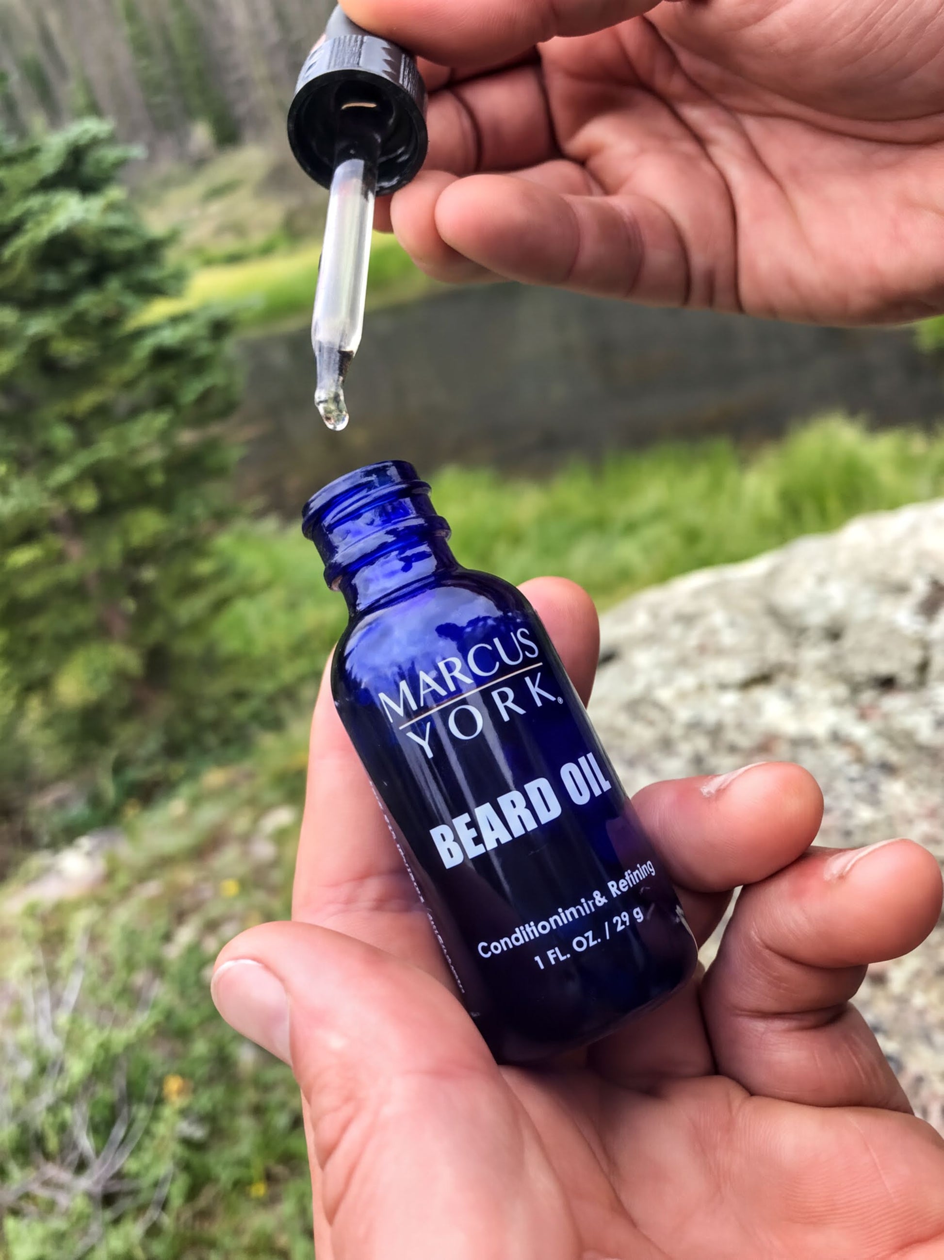 A person's hand holding a blue glass bottle of Marcus York beard oil, with a dropper attached to the bottle, against a natural outdoor background.