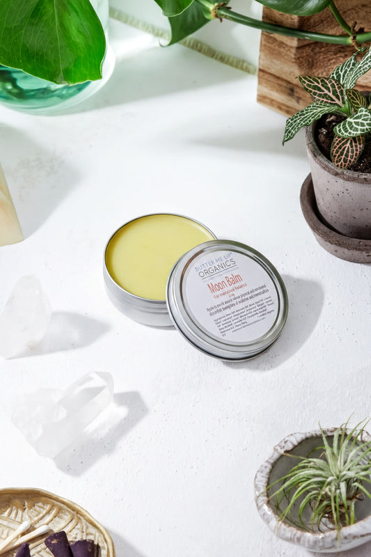 a round tin of "Moisture Balm" with a silver lid, placed on a white surface surrounded by various potted plants and crystals.