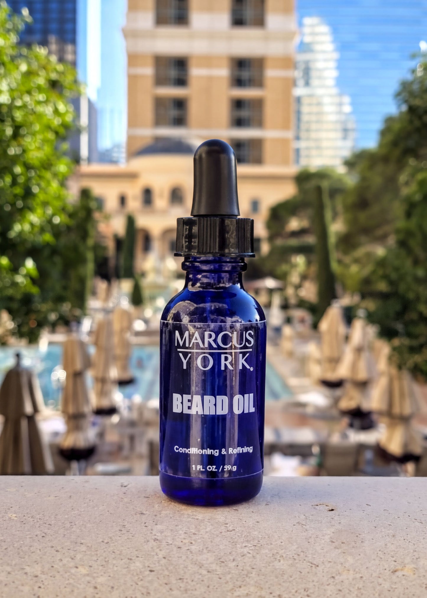 A blue bottle of Marcus York Beard Oil sits on a ledge, with a cityscape and trees visible in the background.