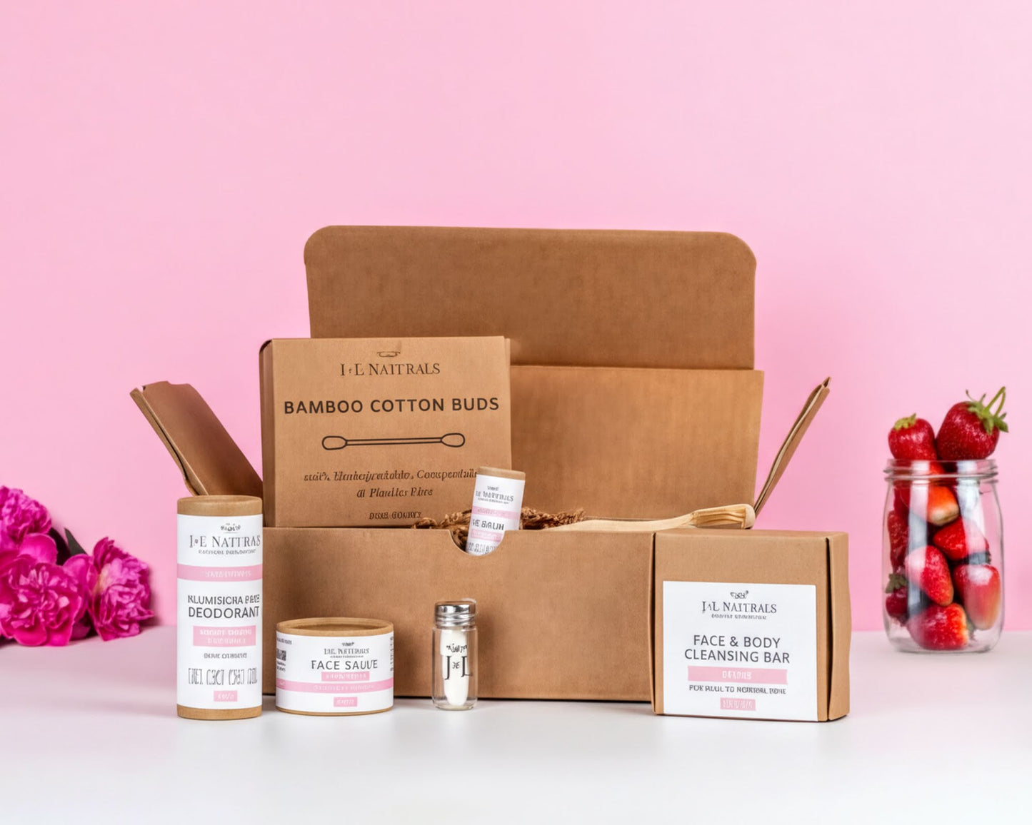 a collection of beauty products from the brand J.L.NATRALS, including a bamboo cotton bud, a face saucer, a face cleanser, and a body cleansing bar, all neatly arranged in a cardboard box. The box is set against a pink background, and there are some strawberries visible in the background as well.