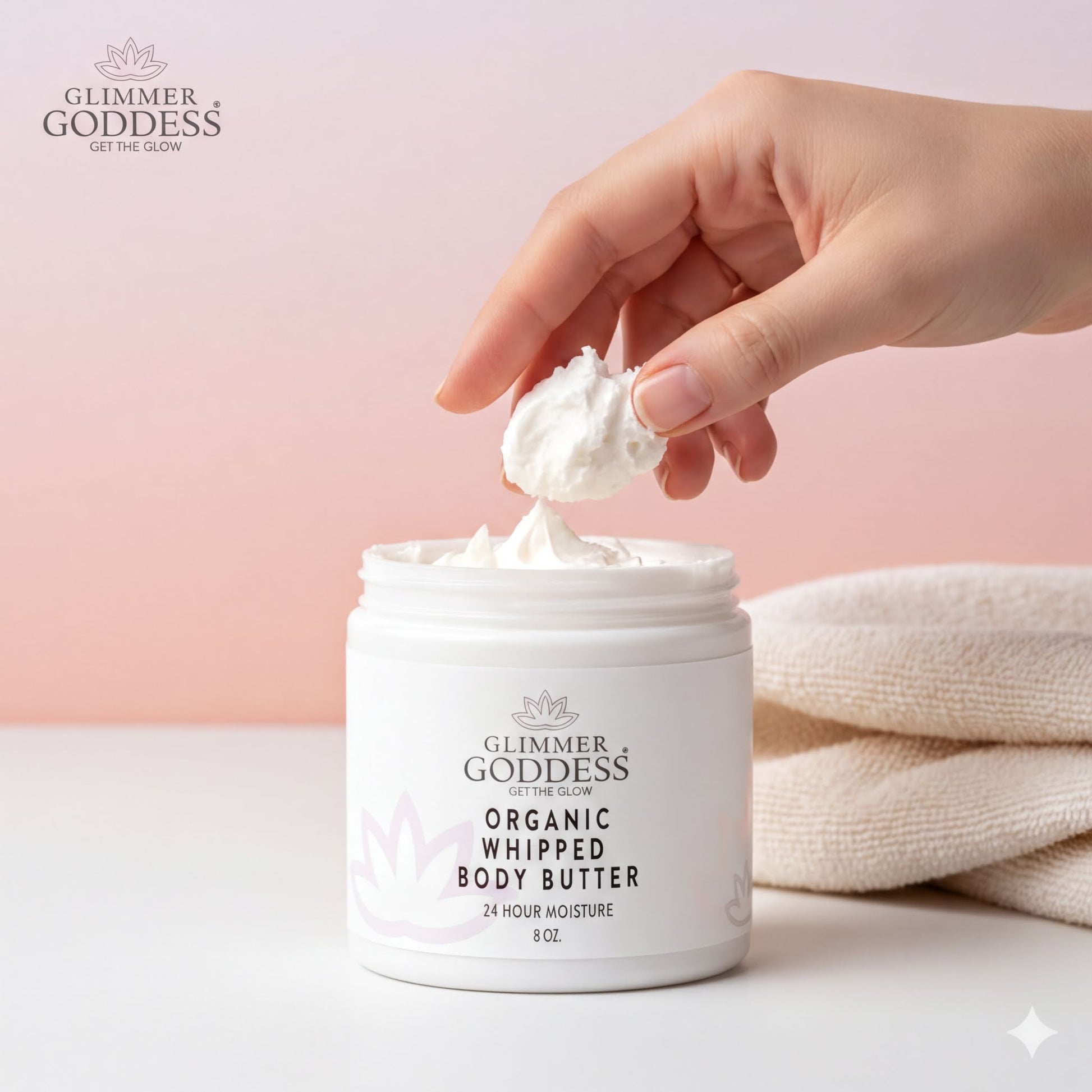 A hand is applying a dollop of organic whipped body butter from a jar labeled "Glimmer Goddess ORGANIC WHIPPED BODY BUTTER 24 HOUR MOISTURE 8 OZ."