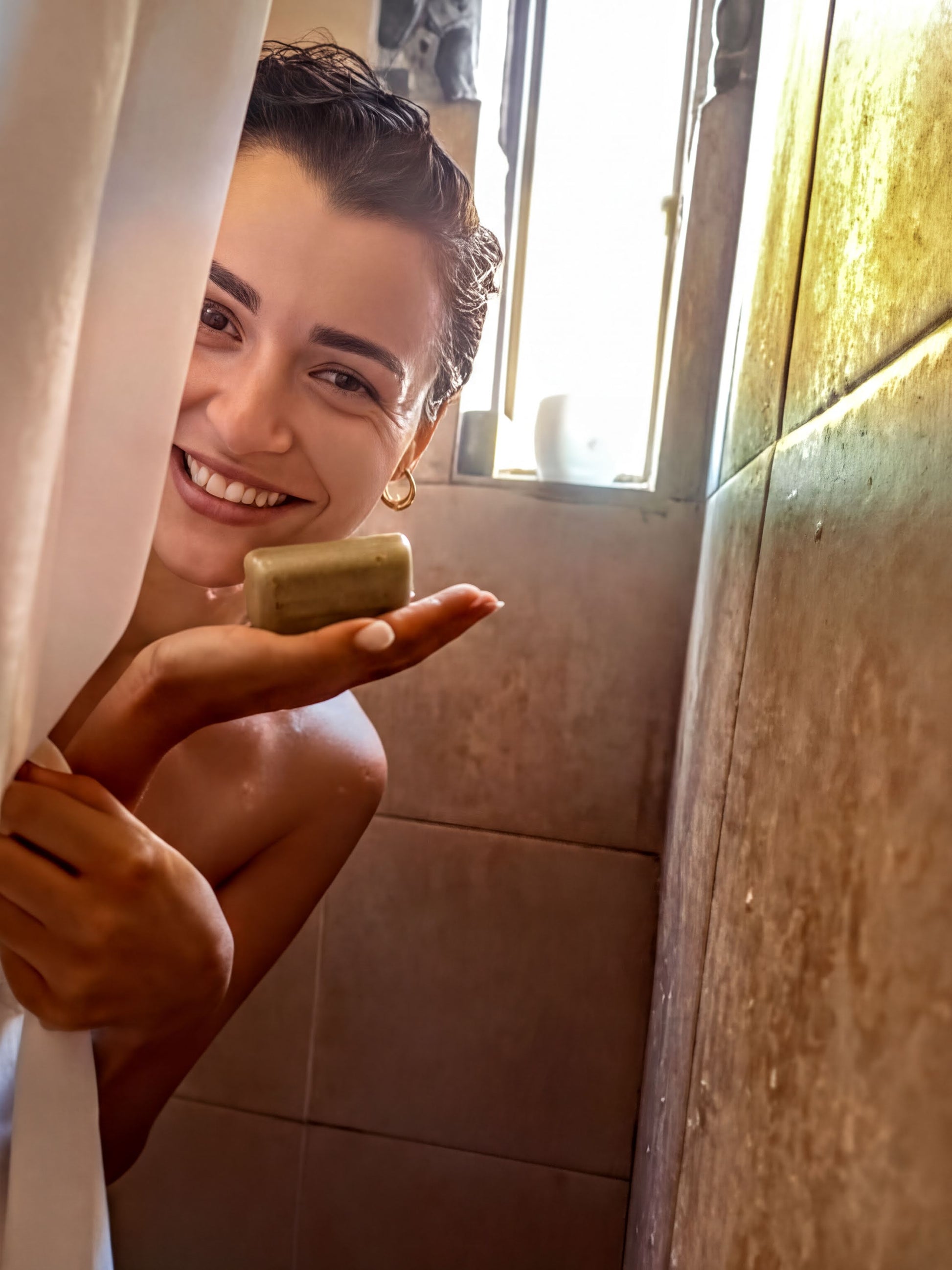 A woman is standing in a bathroom, holding a bar of soap in her hand and smiling.